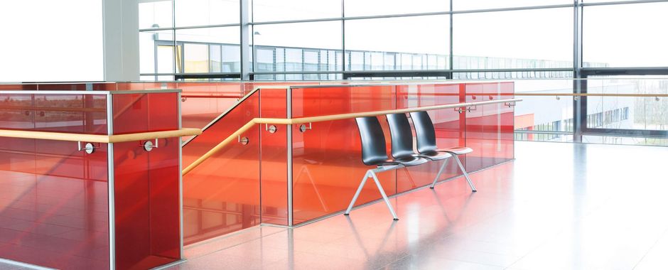 Powder coated red glass partitions in a staircase railing in a well lighted building with three black plastic seats right beside the end of the staircase