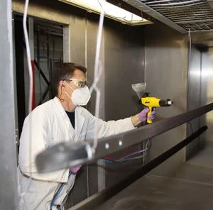 Ein Mann beschichtet schwarze Dachträger in einer Pulverbeschichtungskabine und trägt dabei einen Laborkittel, eine Atemmaske und eine Schutzbrille
