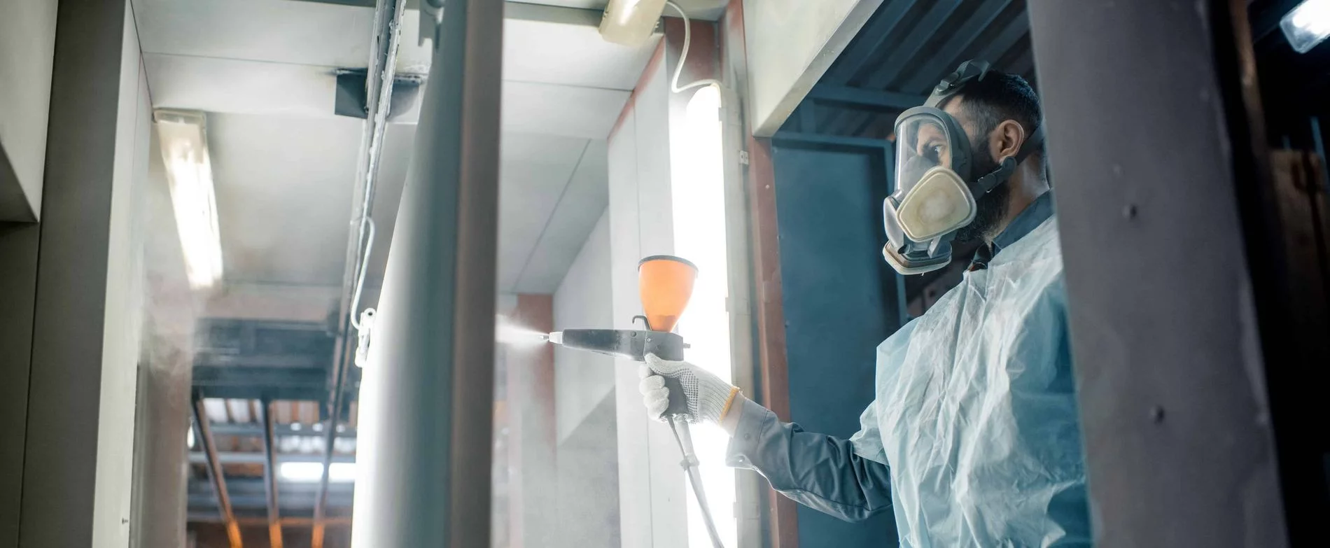 Man wearing a protective suit and respiratory mask is powder coating a metal sheet in a spray booth holding a powder gun in his right hand