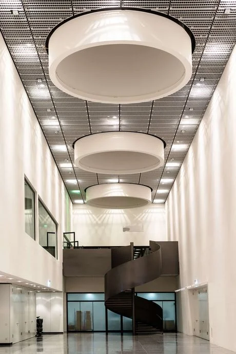 Long haalway with white walls and a black staircase at the end, decorated with 3 large, round, light screens at the ceiling