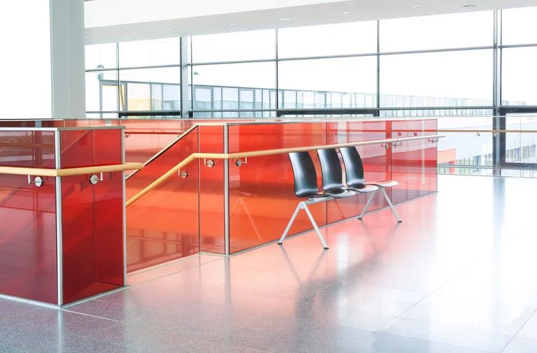 Powder coated red glass partitions in a staircase railing in a well lighted building with three black plastic seats right beside the end of the staircase