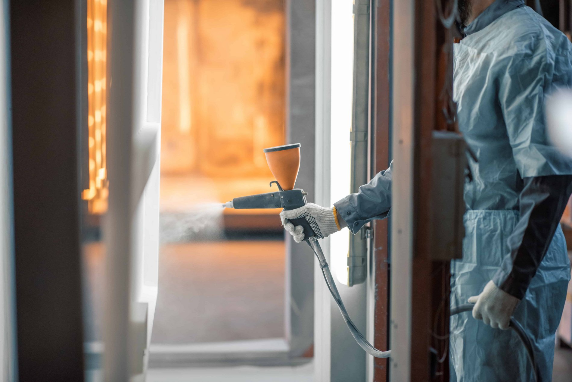 Ein Mann mit Schutzanzug hält eine Pulverpistole in der rechten Hand, während er in einer Spritzkabine ein Metallblech pulverbeschichtet.